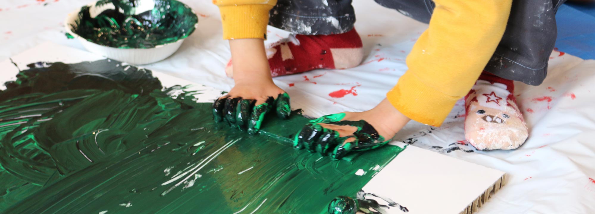 分厚いダンボールに緑色の絵の具を素手で塗り広げている子どもの手元が写っている写真